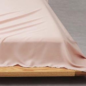 Puede incluir: Una s&aacute;bana de color rosa claro drapeada sobre un marco de cama de madera. La tela tiene una textura suave y lisa con pliegues y arrugas suaves en la superficie, mostrando un color pastel s&oacute;lido que cubre la parte superior y lateral.