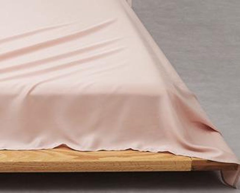 Puede incluir: Una s&aacute;bana de color rosa claro drapeada sobre un marco de cama de madera. La tela tiene una textura suave y lisa con pliegues y arrugas suaves en la superficie, mostrando un color pastel s&oacute;lido que cubre la parte superior y lateral.