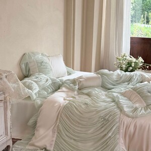 May include: A mint green and white bedding set featuring a ruffled duvet cover with small decorative bows and pearl accents. The set includes matching ruffled pillowcases and a smooth white flat sheet, creating a layered, textured appearance.