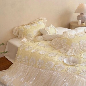 May include: A pale yellow bedding set featuring a textured floral pattern with white daisy-like appliqu&eacute;s. The duvet cover and pillow shams are finished with ruffled edges and a wide, semi-sheer white pleated skirt along the bottom border.