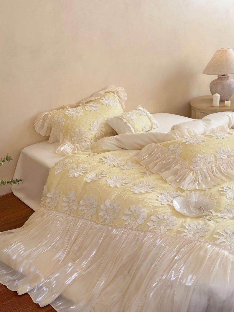 May include: A pale yellow bedding set featuring a textured floral pattern with white daisy-like appliqu&eacute;s. The duvet cover and pillow shams are finished with ruffled edges and a wide, semi-sheer white pleated skirt along the bottom border.