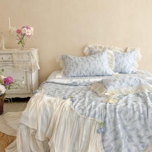 May include: A bed set featuring a light blue quilt and matching pillowcases with a subtle floral pattern. The bedding is accented with cream-colored ruffled fabric trim along the edges. A small white teacup and saucer rest on the bed surface.