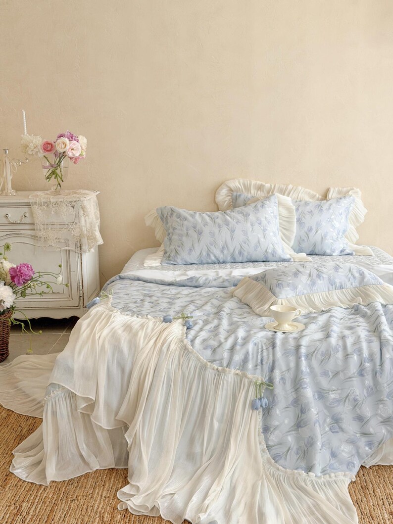 May include: A bed set featuring a light blue quilt and matching pillowcases with a subtle floral pattern. The bedding is accented with cream-colored ruffled fabric trim along the edges. A small white teacup and saucer rest on the bed surface.