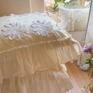 May include: A square cream-coloured throw pillow featuring white embroidered daisy-like floral appliqu&eacute;s and delicate swirling vine patterns. The pillow is finished with two layers of sheer, ruffled fabric trim along the bottom edge.