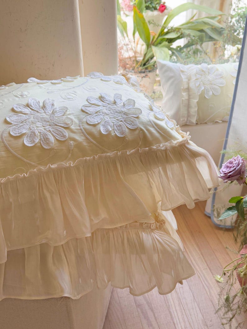 May include: A square cream-coloured throw pillow featuring white embroidered daisy-like floral appliqu&eacute;s and delicate swirling vine patterns. The pillow is finished with two layers of sheer, ruffled fabric trim along the bottom edge.