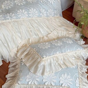 May include: A light blue bedding set featuring a white floral applique pattern with raised petals. The duvet cover and matching rectangular pillows are finished with wide, gathered cream-colored ruffles that have a subtle, glossy sheen.