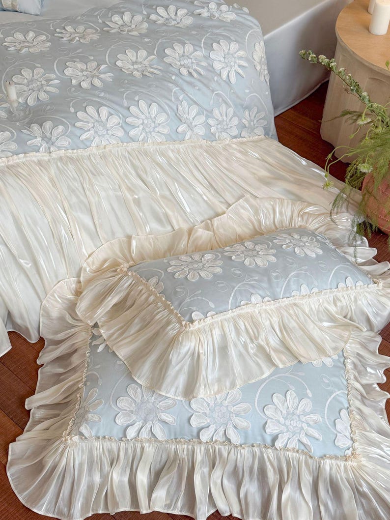 May include: A light blue bedding set featuring a white floral applique pattern with raised petals. The duvet cover and matching rectangular pillows are finished with wide, gathered cream-colored ruffles that have a subtle, glossy sheen.