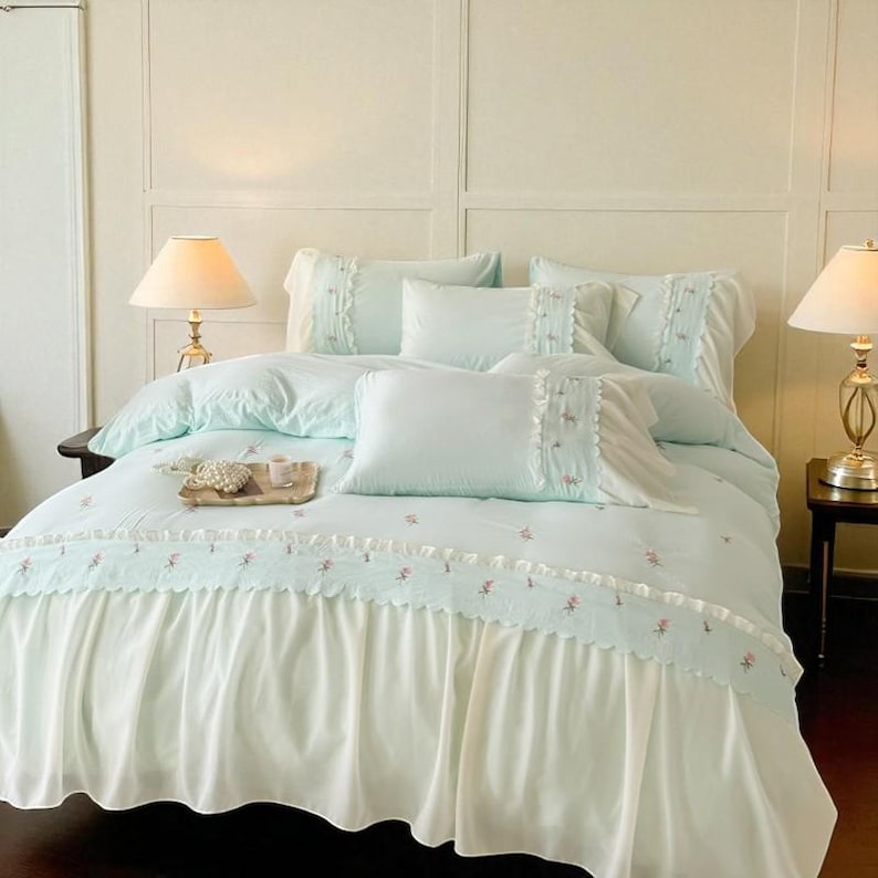 May include: A light blue bed set with a white ruffled bed skirt. The bedding includes a comforter, pillows, and a decorative tray. The comforter and pillowcases have floral embroidery.