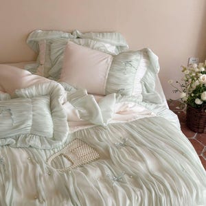 May include: A bedding set featuring a light mint green duvet cover and matching pillowcases with ruffled edges, small fabric bows, and scattered faux pearl accents. A small white beaded handbag rests on top of the textured, pleated fabric surface.