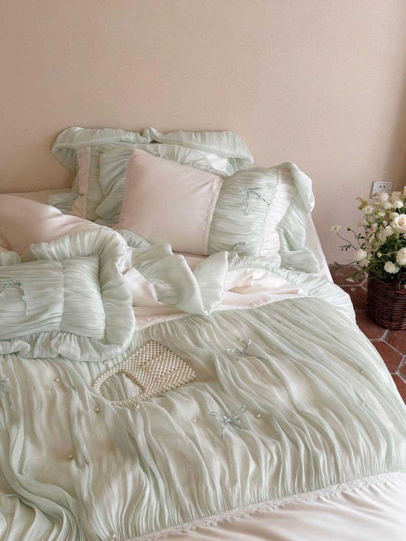 May include: A bedding set featuring a light mint green duvet cover and matching pillowcases with ruffled edges, small fabric bows, and scattered faux pearl accents. A small white beaded handbag rests on top of the textured, pleated fabric surface.