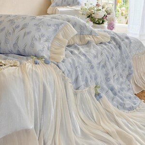 May include: A light blue bedding set featuring a repeating pattern of soft purple tulips. The duvet cover and pillowcases are adorned with cream-colored ruffled fabric edges and small decorative blue tulip accents along the draped skirt.