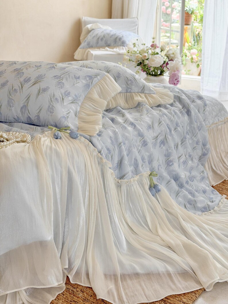 May include: A light blue bedding set featuring a repeating pattern of soft purple tulips. The duvet cover and pillowcases are adorned with cream-colored ruffled fabric edges and small decorative blue tulip accents along the draped skirt.
