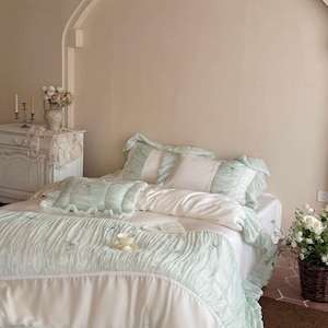 May include: A bedding set featuring a white duvet cover and matching pillowcases with pale mint green ruffled borders and pleated fabric accents. The set includes a rectangular throw pillow and a large coverlet with small decorative bows.