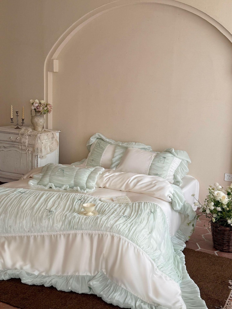May include: A bedding set featuring a white duvet cover and matching pillowcases with pale mint green ruffled borders and pleated fabric accents. The set includes a rectangular throw pillow and a large coverlet with small decorative bows.