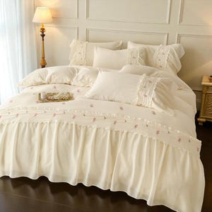 May include: Cream-colored bedding set with embroidered pink floral accents. The set includes a comforter, pillows, and a bed skirt with a ruffled edge. A tray with pearls sits on the comforter. The room has a classic, elegant style.