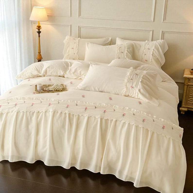 May include: Cream-colored bedding set with embroidered pink floral accents. The set includes a comforter, pillows, and a bed skirt with a ruffled edge. A tray with pearls sits on the comforter. The room has a classic, elegant style.