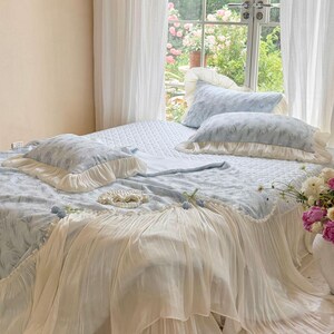 May include: A light blue bedding set featuring a quilted coverlet and two matching pillows, all adorned with a delicate feather-like pattern. The set includes cream-colored ruffled fabric trim along the edges of the pillows and the coverlet.