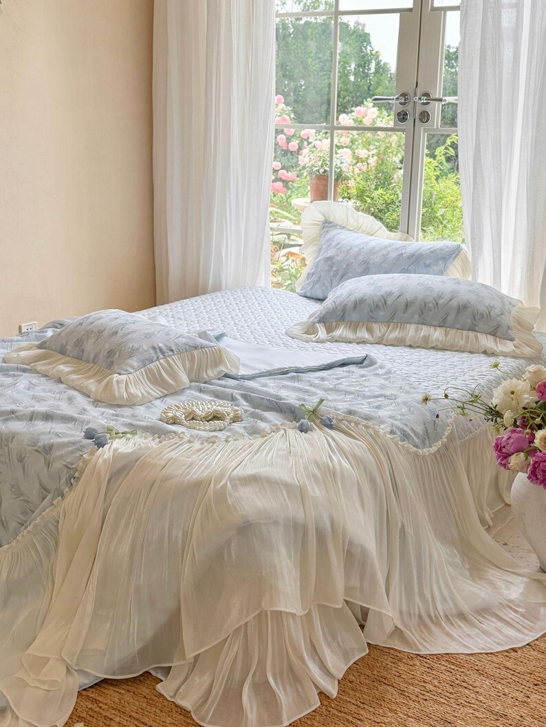 May include: A light blue bedding set featuring a quilted coverlet and two matching pillows, all adorned with a delicate feather-like pattern. The set includes cream-colored ruffled fabric trim along the edges of the pillows and the coverlet.