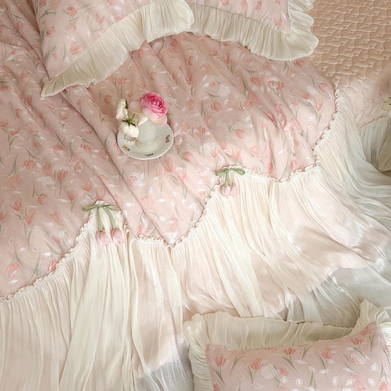 Puede incluir: Un juego de cama rosa claro con un patr&oacute;n de tulipanes repetitivo, adornado con volantes blancos y detalles decorativos de tulipanes de tela. Una peque&ntilde;a taza de t&eacute; de cer&aacute;mica blanca con una flor rosa descansa sobre la tela acolchada.