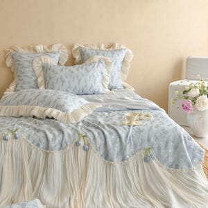 May include: A light blue bedding set featuring a floral pattern, including a quilt with a long, pleated cream-colored ruffle trim adorned with small blue tulip accents, matching pillow shams with ruffled edges, and a coordinating throw pillow.