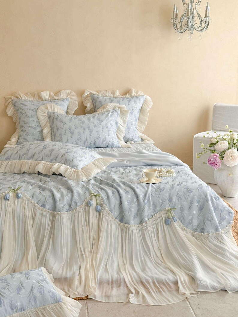 May include: A light blue bedding set featuring a floral pattern, including a quilt with a long, pleated cream-colored ruffle trim adorned with small blue tulip accents, matching pillow shams with ruffled edges, and a coordinating throw pillow.