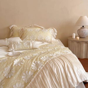 May include: A bedding set featuring a pale yellow duvet cover and matching pillowcases adorned with white floral appliques. The duvet cover includes a wide, pleated, cream-coloured fabric ruffle along the edge, creating a layered texture.
