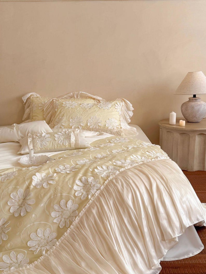 May include: A bedding set featuring a pale yellow duvet cover and matching pillowcases adorned with white floral appliques. The duvet cover includes a wide, pleated, cream-coloured fabric ruffle along the edge, creating a layered texture.