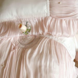 May include: A bedding set featuring a smooth white center panel bordered by pleated pale pink fabric. The design includes delicate white lace trim, small satin bows, and scattered faux pearl embellishments. A clear glass holds a single peach rose.