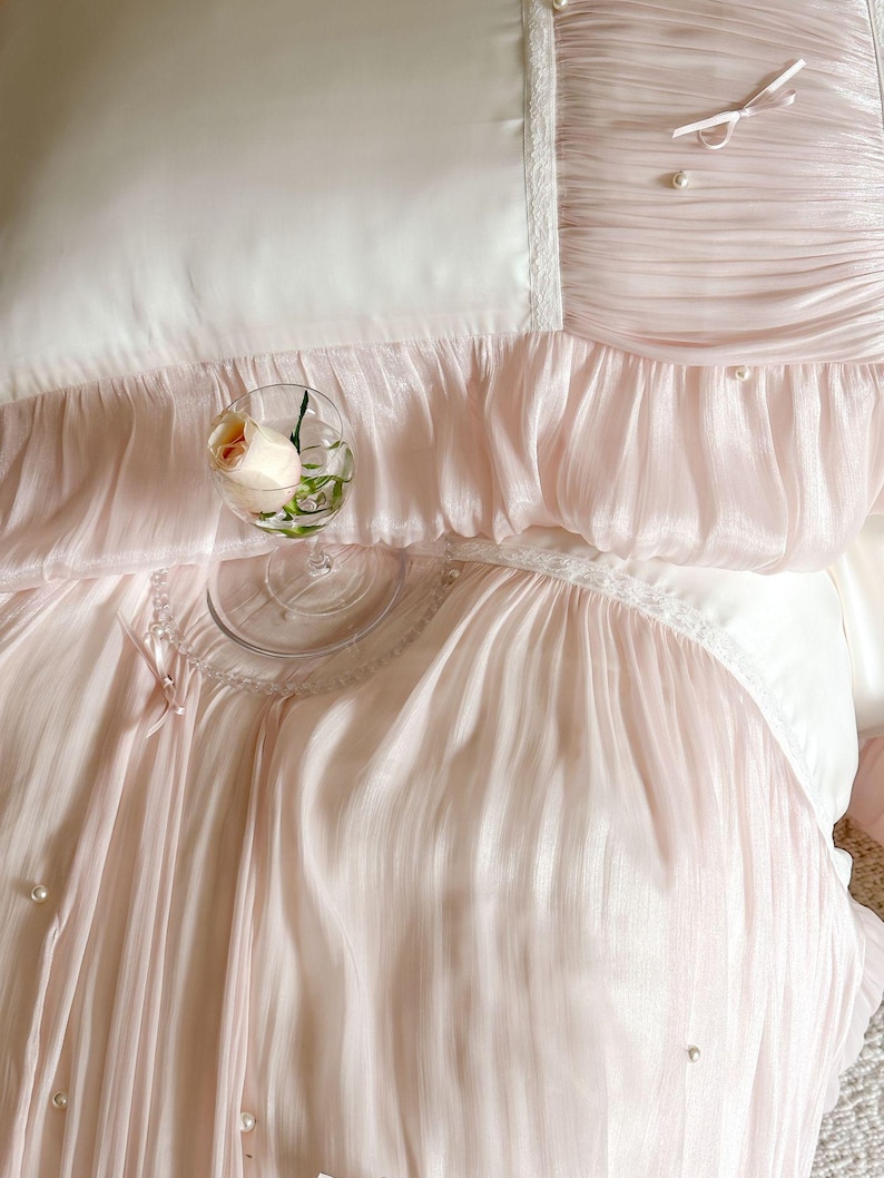 May include: A bedding set featuring a smooth white center panel bordered by pleated pale pink fabric. The design includes delicate white lace trim, small satin bows, and scattered faux pearl embellishments. A clear glass holds a single peach rose.