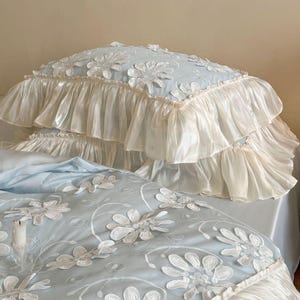 May include: A light blue bedding set featuring a duvet cover and pillowcase with white floral applique embroidery. The edges are finished with wide, gathered cream-colored ruffles that create a layered, voluminous appearance on the fabric.