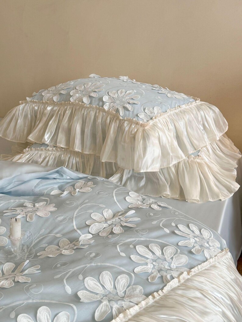 May include: A light blue bedding set featuring a duvet cover and pillowcase with white floral applique embroidery. The edges are finished with wide, gathered cream-colored ruffles that create a layered, voluminous appearance on the fabric.