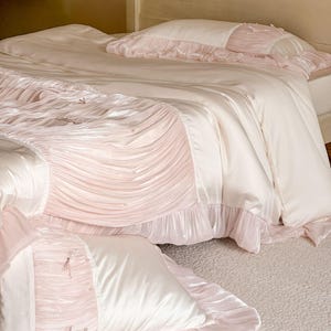 May include: A bedding set featuring a smooth cream-colored duvet cover and matching pillowcases with soft pink pleated fabric panels. The design includes delicate pink ribbon bows and small pearl-like accents, finished with ruffled edges.