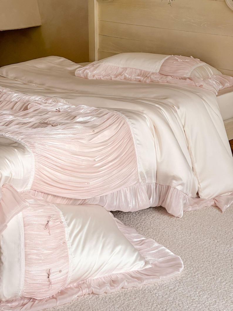 May include: A bedding set featuring a smooth cream-colored duvet cover and matching pillowcases with soft pink pleated fabric panels. The design includes delicate pink ribbon bows and small pearl-like accents, finished with ruffled edges.