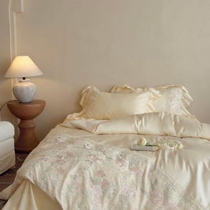 May include: A cream-colored bedding set featuring a duvet cover and two ruffled pillowcases. The duvet cover is adorned with a wide, intricate lace panel displaying a delicate floral pattern in soft pink and green hues, complemented by a string of pearls.