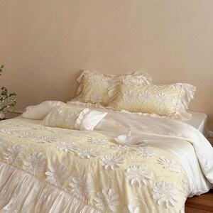 May include: A pale yellow bedding set featuring a textured floral pattern with white raised daisy-like appliqu&eacute;s. The set includes a ruffled duvet cover, two matching ruffled pillow shams, and a smaller rectangular accent pillow with ruffles.