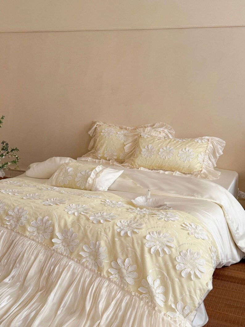 May include: A pale yellow bedding set featuring a textured floral pattern with white raised daisy-like appliqu&eacute;s. The set includes a ruffled duvet cover, two matching ruffled pillow shams, and a smaller rectangular accent pillow with ruffles.