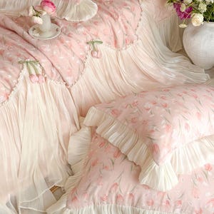 Puede incluir: Un juego de cama rosa claro con un patr&oacute;n repetitivo de tulipanes y ribetes con volantes de color crema. El conjunto incluye una colcha acolchada y fundas de almohada a juego, adornadas con delicados tulipanes de tela en los bordes.