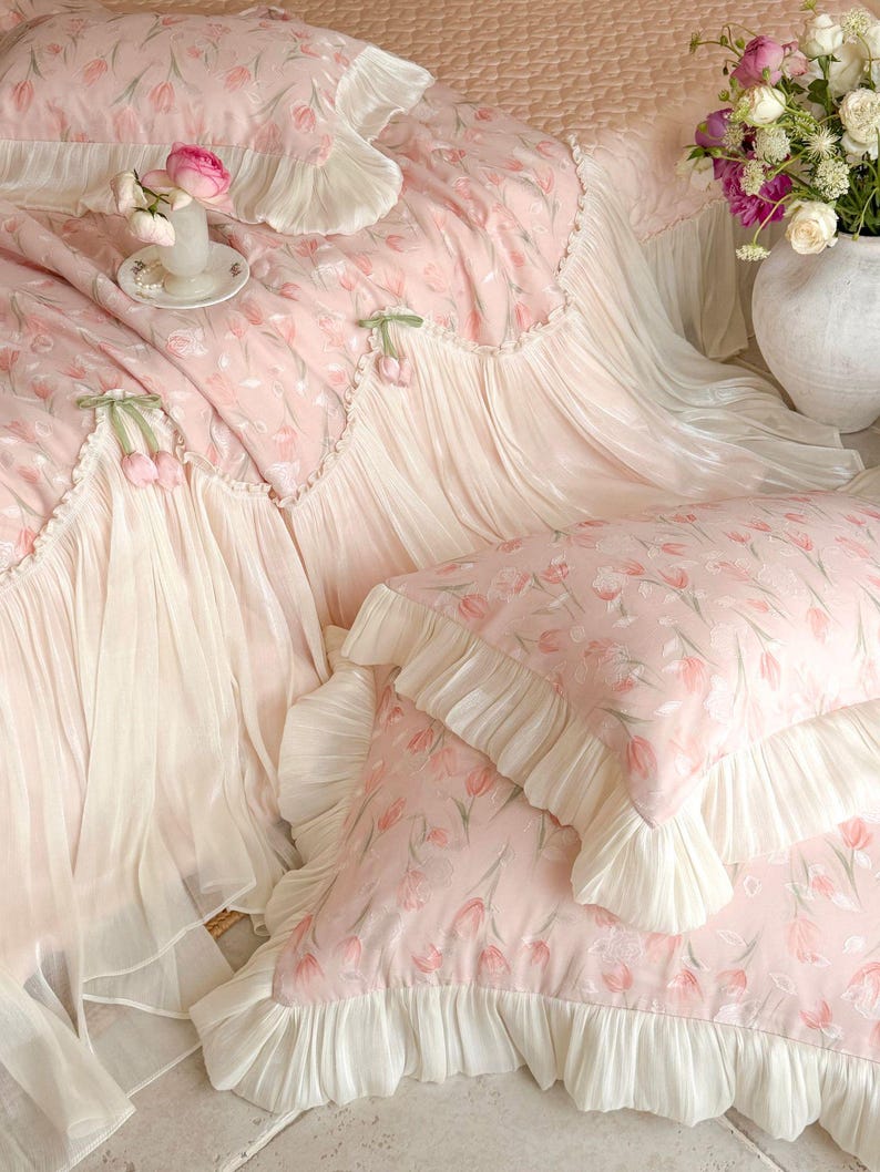 Puede incluir: Un juego de cama rosa claro con un patr&oacute;n repetitivo de tulipanes y ribetes con volantes de color crema. El conjunto incluye una colcha acolchada y fundas de almohada a juego, adornadas con delicados tulipanes de tela en los bordes.