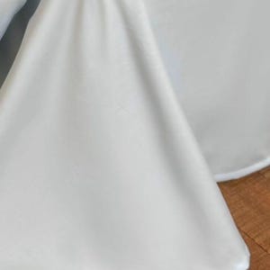 May include: A solid white fabric drape featuring a smooth, matte texture and a clean, finished hem along the bottom edge. The material hangs in soft, vertical folds, creating a series of gentle curves and shadows across the surface of the cloth.