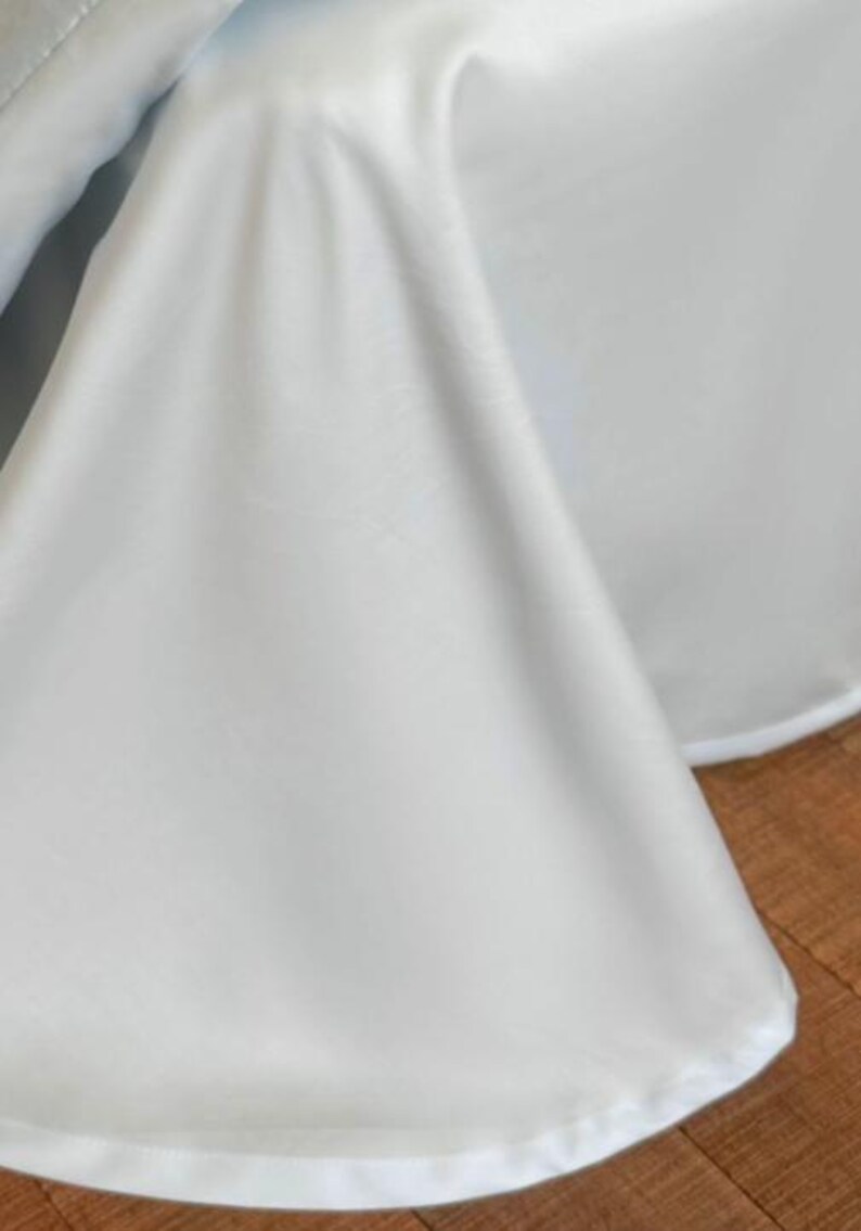 May include: A solid white fabric drape featuring a smooth, matte texture and a clean, finished hem along the bottom edge. The material hangs in soft, vertical folds, creating a series of gentle curves and shadows across the surface of the cloth.