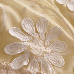 May include: A pale yellow fabric featuring large, white, embroidered floral motifs with detailed petal stitching. The textile has a soft, gathered, and ruffled border at the bottom edge, creating a textured appearance with layered, delicate, and light-coloured floral patterns.