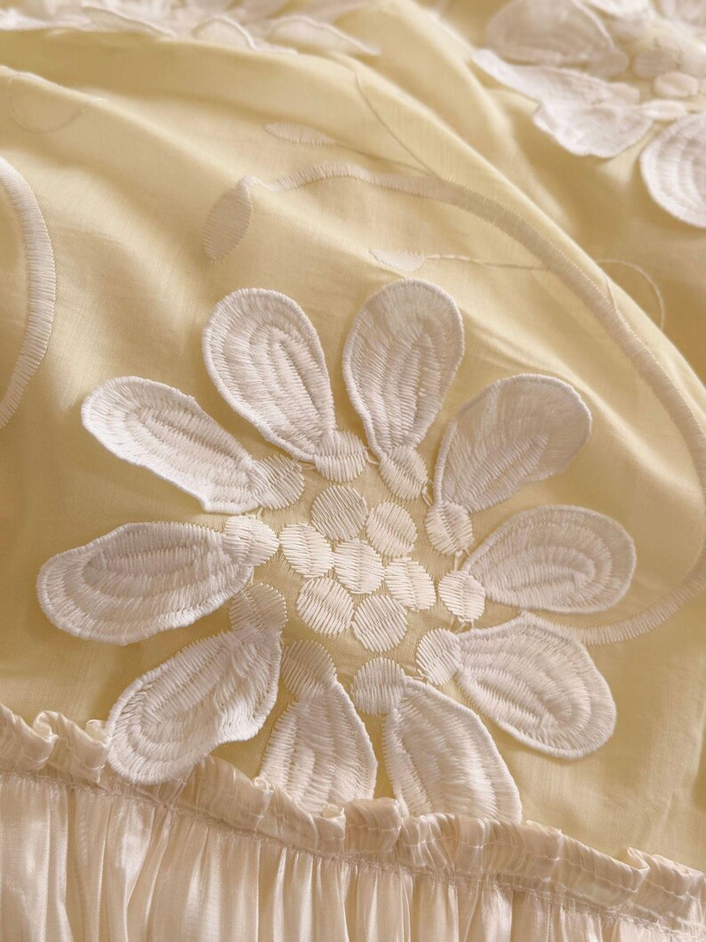 May include: A pale yellow fabric featuring large, white, embroidered floral motifs with detailed petal stitching. The textile has a soft, gathered, and ruffled border at the bottom edge, creating a textured appearance with layered, delicate, and light-coloured floral patterns.