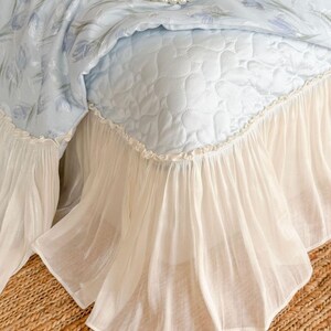 May include: A light blue bedspread featuring a quilted heart pattern and a floral print top layer. The bedspread is finished with a gathered, sheer cream-colored ruffled skirt along the bottom edge. A string of white faux pearls rests on top.