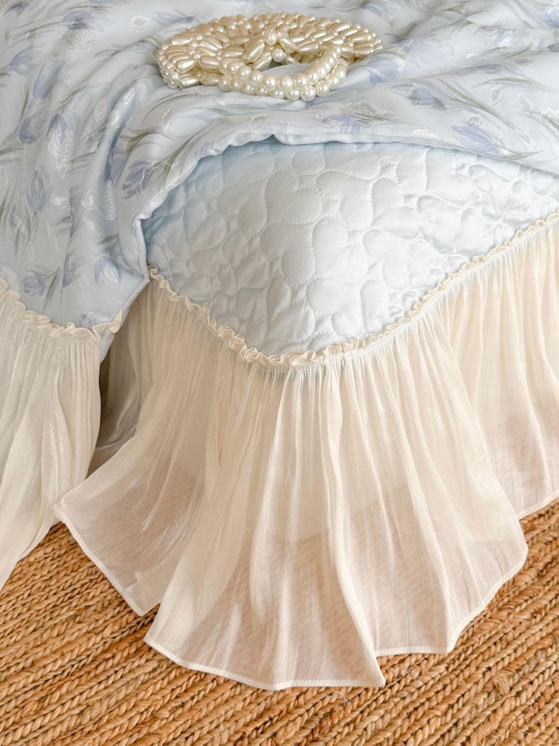 May include: A light blue bedspread featuring a quilted heart pattern and a floral print top layer. The bedspread is finished with a gathered, sheer cream-colored ruffled skirt along the bottom edge. A string of white faux pearls rests on top.