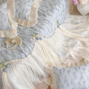 May include: A light blue bedding set featuring a floral pattern, cream-colored ruffled edges, and decorative fabric tulip accents. The set includes a quilted coverlet and matching rectangular pillows, all finished with delicate pleated trim.