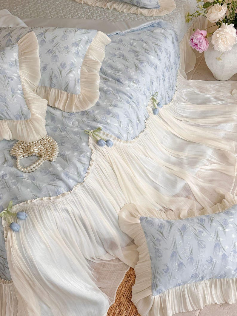 May include: A light blue bedding set featuring a floral pattern, cream-colored ruffled edges, and decorative fabric tulip accents. The set includes a quilted coverlet and matching rectangular pillows, all finished with delicate pleated trim.