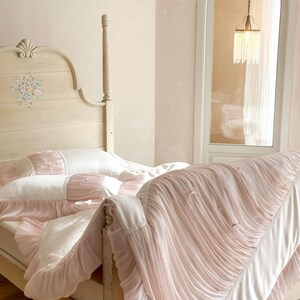 May include: A cream-colored wooden bed frame with a floral headboard design, featuring white and pale pink bedding. The duvet and pillowcases are adorned with gathered fabric ruffles, small white bows, and tiny pearl-like accents throughout.