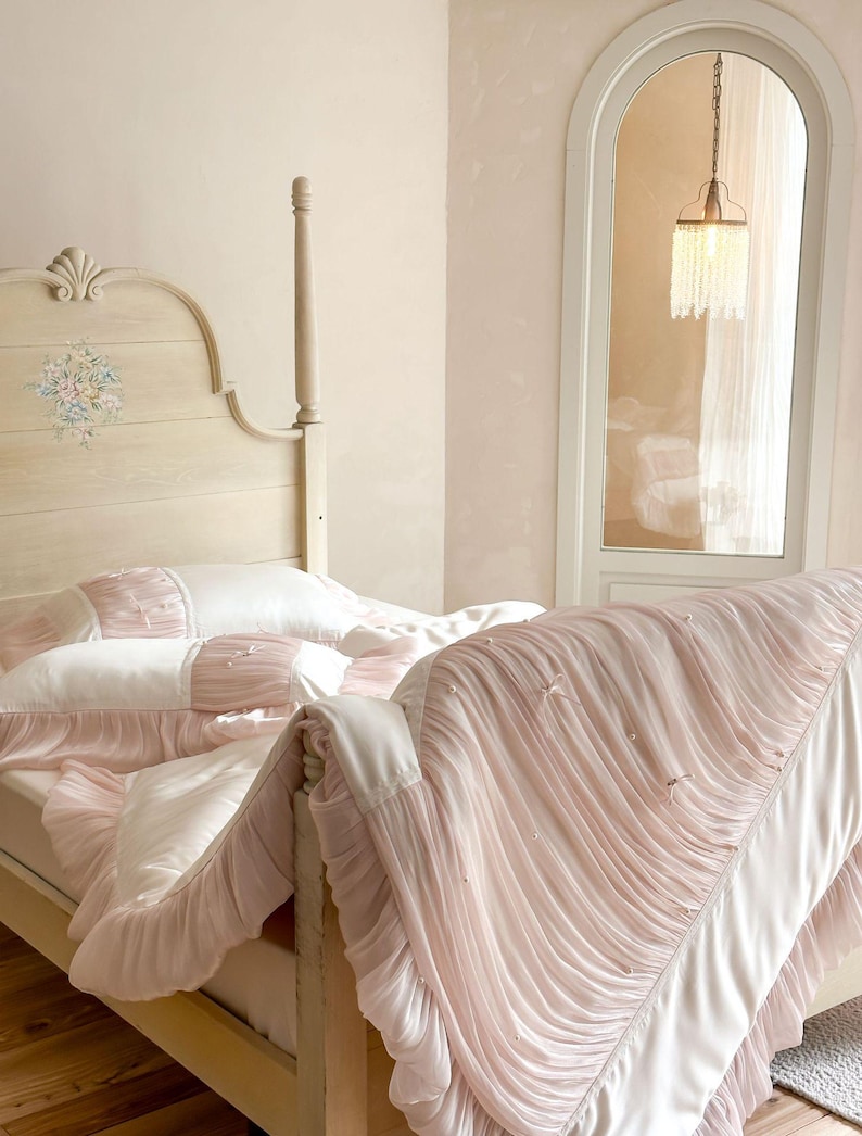 May include: A cream-colored wooden bed frame with a floral headboard design, featuring white and pale pink bedding. The duvet and pillowcases are adorned with gathered fabric ruffles, small white bows, and tiny pearl-like accents throughout.