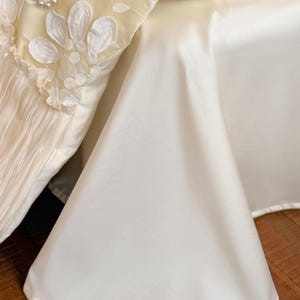 May include: A cream-coloured bed valance featuring a pleated section with white floral embroidery and a string of faux pearls draped across the top. The fabric has a smooth, satin-like finish and a clean, hemmed edge that drapes towards the floor.