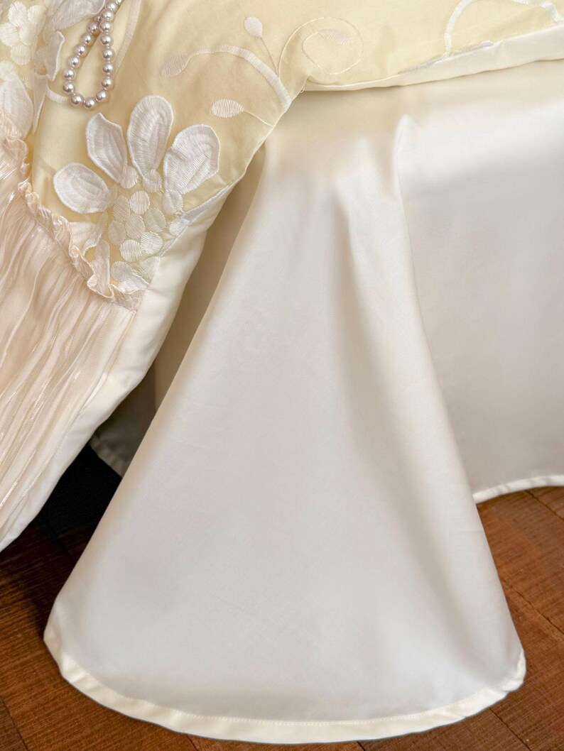 May include: A cream-coloured bed valance featuring a pleated section with white floral embroidery and a string of faux pearls draped across the top. The fabric has a smooth, satin-like finish and a clean, hemmed edge that drapes towards the floor.
